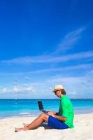 junger Mann, der am Laptop am tropischen Strand arbeitet foto