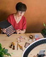 Frau mittleren Alters, die über Kosmetik mit Make-up-Lidschatten und Rouge-Palette und -Pinseln spricht, während sie ihr Video aufnimmt. reife Frau, die ein Video für ihren Blog über Kosmetik macht foto