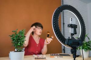 Beauty-Bloggerin oder Influencerin mittleren Alters mit Make-up-Produktaufnahmevideo im Zimmer zu Hause. mit Ringlampe und Smartphone foto