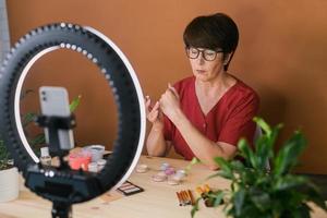 Frau mittleren Alters, die über Kosmetik mit Make-up-Lidschatten und Rouge-Palette und -Pinseln spricht, während sie ihr Video aufnimmt. reife Frau, die ein Video für ihren Blog über Kosmetik macht foto