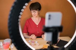 Frau mittleren Alters, die über Kosmetik mit Make-up-Lidschatten und Rouge-Palette und -Pinseln spricht, während sie ihr Video aufnimmt. reife Frau, die ein Video für ihren Blog über Kosmetik macht foto