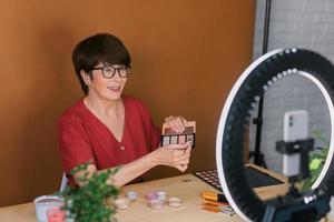 Beauty-Bloggerin oder Influencerin mittleren Alters mit Make-up-Produktaufnahmevideo im Zimmer zu Hause. mit Ringlampe und Smartphone foto