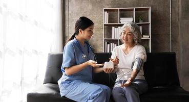 glückliche Seniorin, die ihren Blutdruck in einem Pflegeheim von ihrer Pflegekraft messen lässt. Glückliche Krankenschwester, die den Blutdruck einer älteren Frau im Wohnzimmer misst