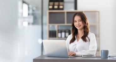 charmante asiatische frau, die im büro arbeitet und einen laptop verwendet, der in die kamera schaut. foto