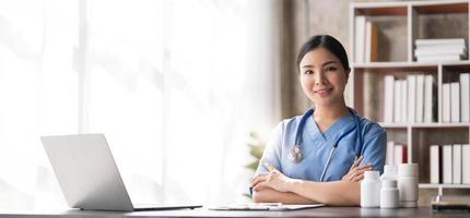 Junge schöne Frau des asiatischen Arztes, die lächelt, indem sie mit einem Laptop-Computer arbeitet und etwas auf Papierkram oder weißes Papier der Zwischenablage im Krankenhaus-Schreibtischbüro schreibt, medizinisches Konzept im Gesundheitswesen foto