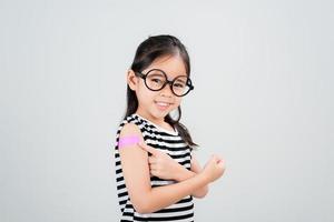 asiatische kleine mädchen tragen eine brille, die ihren arm mit verband nach der virusimpfstoffimpfung zeigt glückliches kind und kinderimpfungskampagne. schützen sie ihr kind mit einem impfkonzept foto