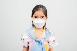 asiatisches kleines mädchen, das eine atemschutzmaske trägt, um den ausbruch des coronavirus zu schützen und die hand auf einen leeren hintergrund zeigt, neues virus covid-19 foto