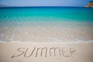 Wort Sommer handschriftlich am Sandstrand mit sanften Meereswellen im Hintergrund foto