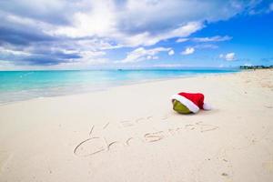 frohe weihnachten geschrieben am weißen sand des strandes mit roter weihnachtsmütze foto