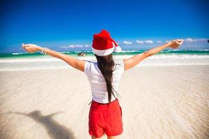 Rückansicht eines schönen Mädchens mit Weihnachtsmütze, das wie ein Vogel am weißen Strand spazieren geht foto