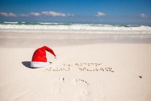 weihnachtsmannmütze am weißen sandstrand und frohes neues jahr in den sand geschrieben foto