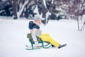 Entzückendes kleines glückliches Mädchen, das im verschneiten Wintertag Rodeln fährt. foto