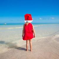 Rückansicht des kleinen süßen Mädchens mit rotem Hut Weihnachtsmann am Strand foto