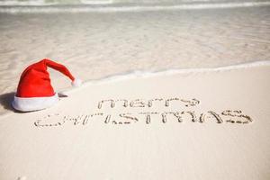 frohe weihnachten geschrieben am weißen sand des tropischen strandes mit weihnachtshut foto
