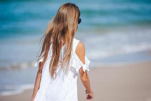 Rückansicht eines entzückenden kleinen Mädchens mit langen Haaren im weißen Kleid, das im tropischen Strandurlaub spazieren geht foto