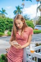 schönes jugendlich Mädchen, das Smartphone verwendet und trinkt, nimmt Getränk im Freien mit foto