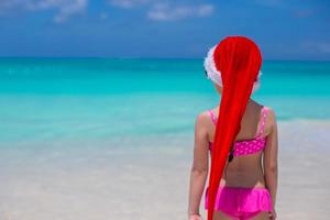 Rückansicht des süßen Mädchens mit rotem Hut Weihnachtsmann am Strand foto