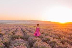 süßes mädchen auf dem lavendelblumenfeld bei sonnenuntergang im purpurroten kleid foto