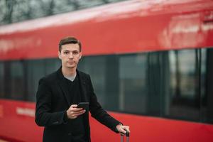 junger kaukasischer mann mit smarphone und gepäck am bahnhof, der mit dem zug reist foto