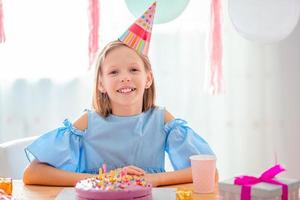 kaukasisches mädchen lächelt träumerisch und betrachtet geburtstagsregenbogenkuchen. festlicher bunter hintergrund mit luftballons. geburtstagsfeier und wunschkonzept. foto