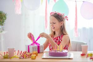 kaukasisches mädchen lächelt träumerisch und betrachtet geburtstagsregenbogenkuchen. festlicher bunter hintergrund mit luftballons. geburtstagsfeier und wunschkonzept. foto