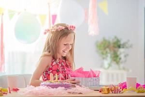 kaukasisches mädchen lächelt träumerisch und betrachtet geburtstagsregenbogenkuchen. festlicher bunter hintergrund mit luftballons. geburtstagsfeier und wunschkonzept. foto