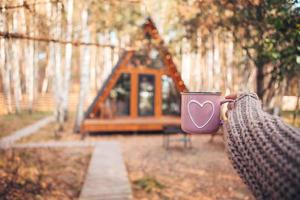 Heiße Tasse Tee, die die Hände der Frau im Wollpullover auf dem Hintergrund des gemütlichen Hauses wärmt foto