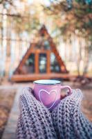 Heiße Tasse Tee, die die Hände der Frau im Wollpullover auf dem Hintergrund des gemütlichen Hauses wärmt foto