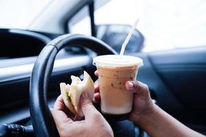 asiatische fahrerin hält eiskaffeetasse und sandwichbrot zum essen und trinken im auto, gefährlich und riskiert einen unfall. foto
