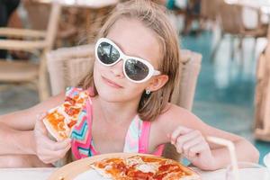 Porträt eines süßen kleinen Mädchens, das am Esstisch sitzt und Pizza isst foto