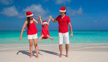 glückliche dreiköpfige familie in weihnachtsmützen, die spaß am weißen strand haben foto