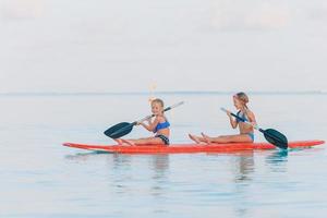 Kleine süße Mädchen, die während der Sommerferien auf dem Surfbrett schwimmen foto