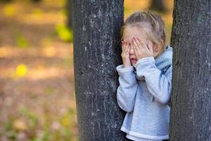 Kleines Mädchen, das Verstecken spielt und in der Nähe des Baums im Herbstpark sucht foto