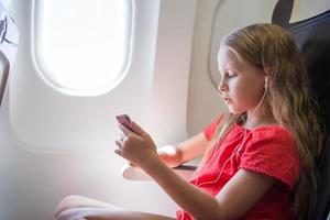 entzückendes kleines Mädchen, das mit einem Flugzeug reist. kind hört musik, die in der nähe des flugzeugfensters sitzt foto