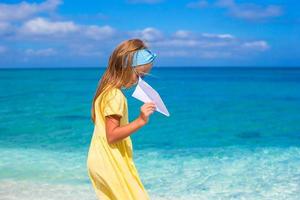 Fröhliches kleines Mädchen spielt mit Papierflugzeug am weißen Sandstrand foto