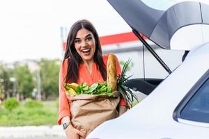 Junge Frau, die einen Einkaufswagen voller Lebensmittel auf dem Parkplatz im Freien reitet. junge Frau auf dem Parkplatz, Laden von Einkäufen in den Kofferraum des Autos. erfolgreich eingekauft. Frau, die nach dem Einkaufen Taschen ins Auto legt foto