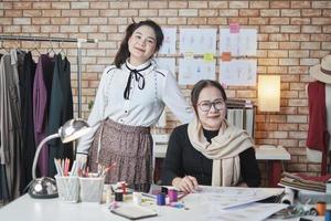 asiatische modedesignerin und junge lehrling schauen in die kamera und lächeln im studio, arme verschränkt, arbeiten mit buntem faden für kleiderdesign, boutique schneider sme unternehmer. foto