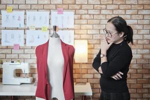 asiatische reife Modedesignerin steht in der Nähe von Ankleidepuppen, denkt nach und erwägt Designideen im Studio, arbeitet mit Kleiderkollektion, professioneller Boutique-Schneider und KMU-Unternehmer. foto