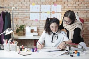 asiatische modedesignerin mittleren alters unterrichtet einen jungen jugendlichen schneider im studio mit buntem faden und nähstoff für kleiderdesign-kollektionsideen, professionelles boutique-kleinunternehmen. foto