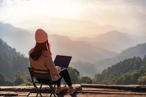 junge freiberufliche reisende, die online mit laptop arbeitet und die wunderschöne naturlandschaft mit bergblick bei sonnenaufgang genießt foto