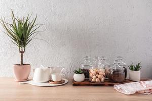 Kücheninnenraum, Hintergrund. Gläser mit trockenem Tee, Keksen, Kaffeebohnen, Teekanne und Tassen aus weißer Keramik. Pflanzen. Vorderansicht.