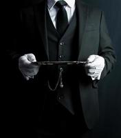 Porträt eines Butlers oder Kellners in einem dunklen Anzug und weißen Handschuhen, die ein silbernes Serviertablett halten. Konzept der Dienstleistungsbranche und tadelloser Gastfreundschaft. foto