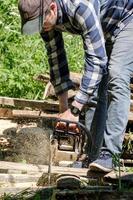 Ein Mann sägt an einem sonnigen Tag alte Bretter für Brennholz mit einer professionellen Kettensäge auf einem Dorfhof. foto