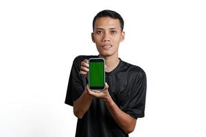 Aufgeregter asiatischer Mann mit schwarzem Trainings-T-Shirt, der auf den grünen Bildschirm des Smartphones zeigt. durch weißen Hintergrund isoliert foto