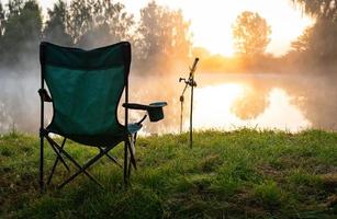 Fischerstuhl und Angelruten im Hintergrund über dem See bei Sonnenaufgang, nebliger Morgen. Angelkonzept. foto