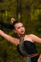 asiatische Frau, die ihr schwarzes Haar in einem grünen Kostüm hält, während sie Make-up trägt und während des Tanzfestivals vor dem Wald posiert foto