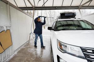 Mann wäscht Hochdruckwasser amerikanisches SUV-Auto mit Dachgepäckträger bei Selbstbedienungswäsche bei kaltem Wetter. foto