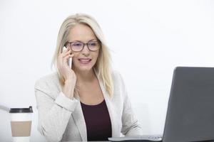 auf die Anforderungen der Kunden hören. Schöne Frau mittleren Alters mit Brille, die mit dem Smartphone spricht und an ihrem Arbeitsplatz im Büro lächelt. foto