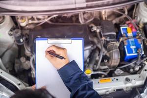 Hände junger professioneller Mechaniker in einheitlicher Schrift auf Klemmbrett gegen Auto in offener Motorhaube in der Werkstatt. Wartung se. foto