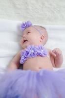 nahaufnahme niedliches neugeborenes baby in lila bodysuit, das sich allein auf dem bett hinlegt. entzückendes kind ruht auf weißen bettlaken und starrt auf die kamera, die friedlich aussieht. Säuglings-, Gesundheitswesen und Pädiatrie, Säuglingskonzept foto